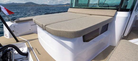 Modern motorboat deck featuring a beige cushioned sun pad on white fiberglass, calm blue sea and distant coastal hills under a partly cloudy sky — perfect for sunbathing.