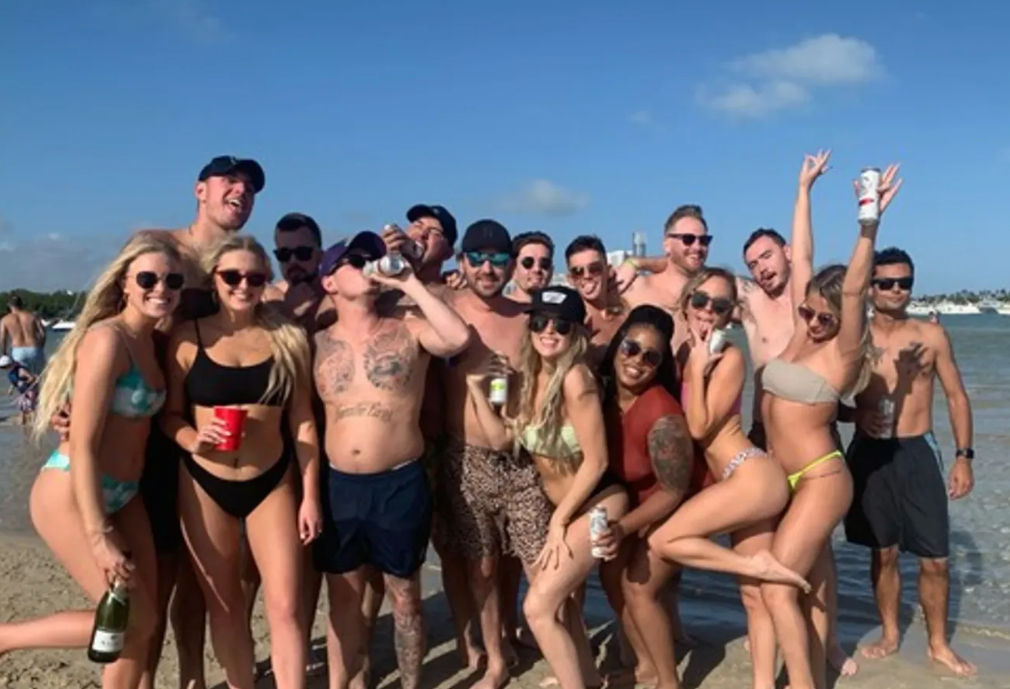 Group of friends in bikinis and swim trunks posing with drinks on a sunny beach shoreline, with boats anchored in the shallow bay behind them.
