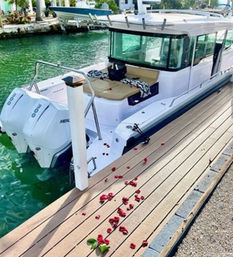 White cabin motorboat with twin outboard engines moored at a turquoise-water marina, tan cockpit cushions and a romantic trail of red rose petals scattered along the wooden dock.