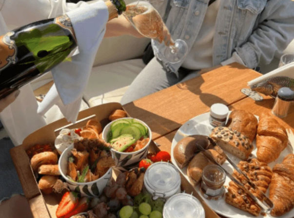 Sunny picnic-style brunch on a wooden table with champagne being poured into a glass, croissants and seeded rolls, bagels, avocado-topped bowls, strawberries, grapes and jarred spreads.