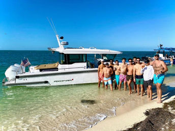 Sunlit group of shirtless men posing on a sandy beach beside a white motorboat beached in clear turquoise water under a bright blue sky