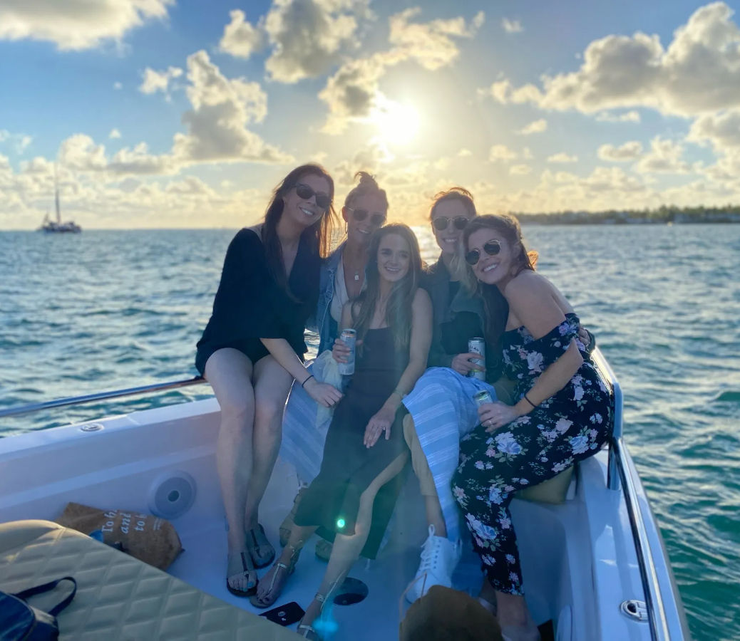 Group of five friends smiling on a small boat at sunset over calm ocean waters, holding drinks with sunlit clouds and a distant sailboat on the horizon.