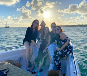 Group of five friends smiling on a small boat at sunset over calm ocean waters, holding drinks with sunlit clouds and a distant sailboat on the horizon.