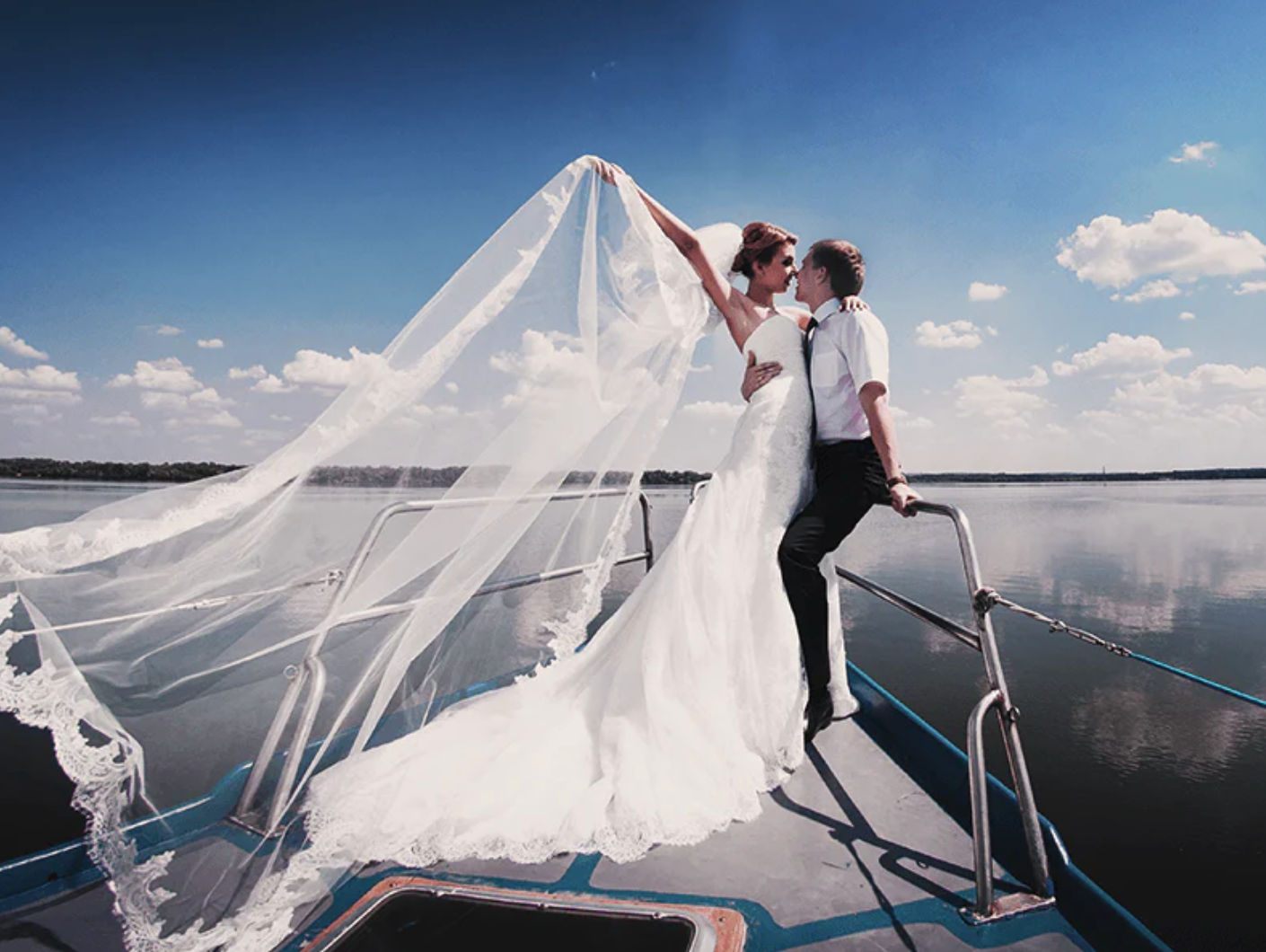 Romantic bride and groom kissing on the bow of a boat on a calm lake, bride’s long white gown and veil streaming in the breeze beneath a bright blue sky with fluffy clouds.