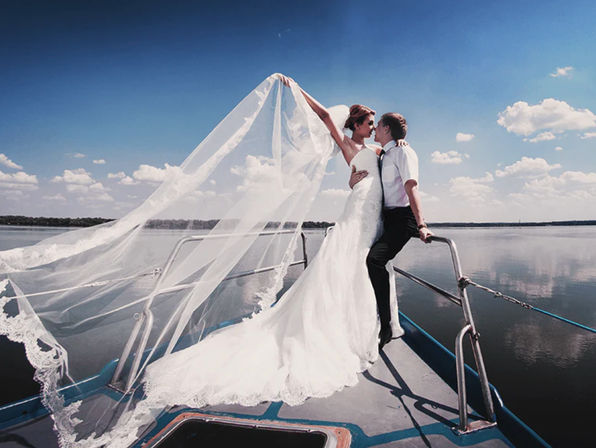 Romantic bride and groom kissing on the bow of a boat on a calm lake, bride’s long white gown and veil streaming in the breeze beneath a bright blue sky with fluffy clouds.