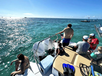 Family boating outing on a sunny day — people lounging on the stern of a motorboat over clear turquoise water, children in life jackets, snorkel fins and a cooler on deck, and an American flag fluttering.