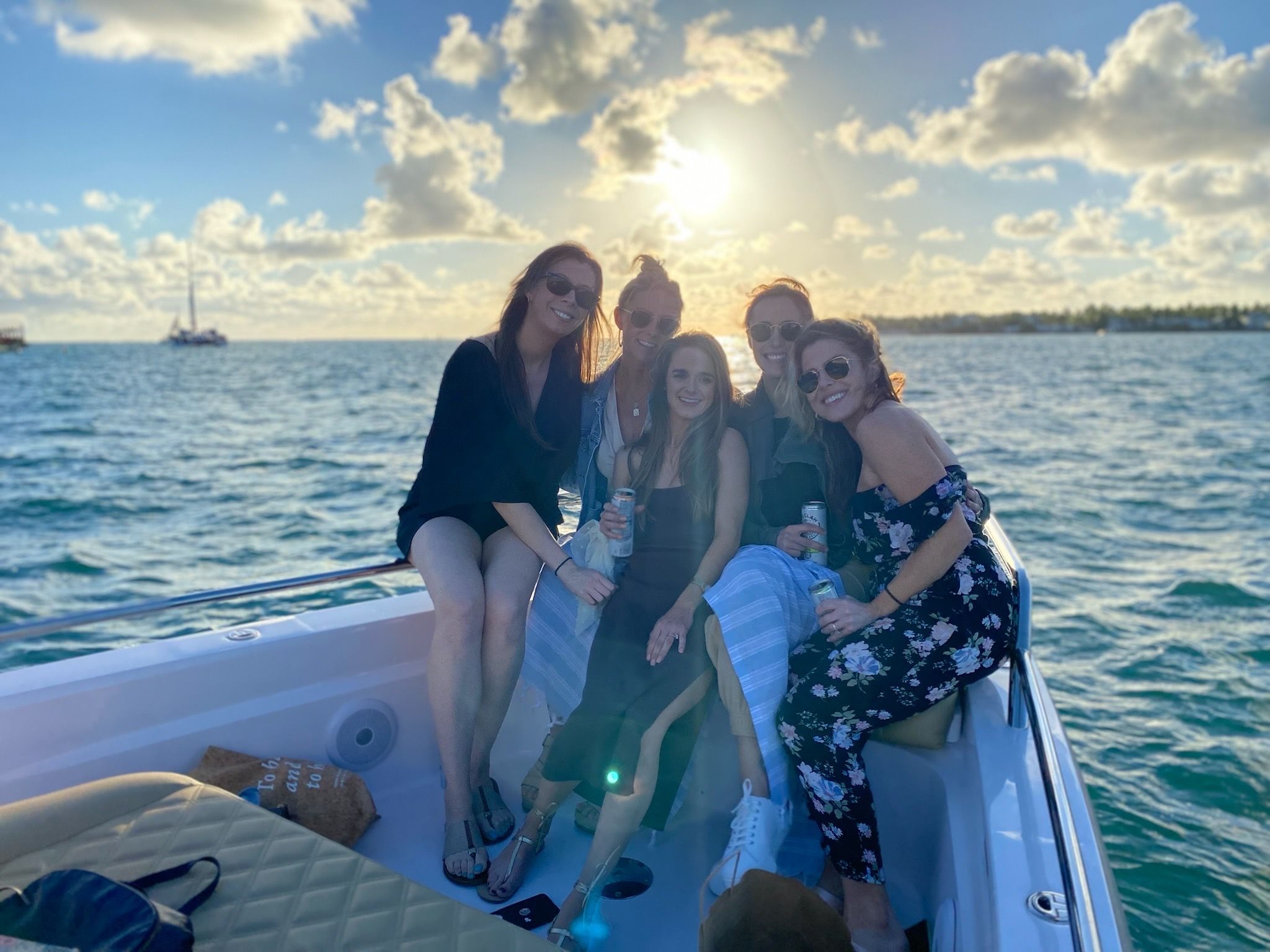 Five friends smiling and holding drinks on a small boat at sunset, sunlit coastal waters and a sailboat on the horizon.