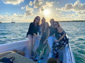 Five friends smiling on a motorboat during a golden ocean sunset, holding drinks with a sailboat on the horizon