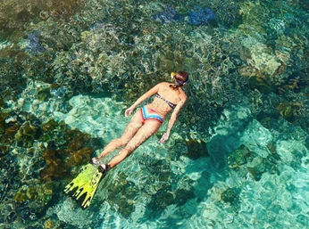 Snorkeler in a colorful bikini and yellow fins floating face-down over crystal-clear turquoise water above a vibrant coral reef