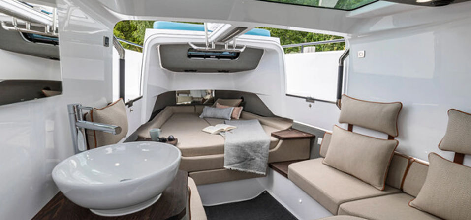 Sunlit modern yacht cabin interior: cozy V-berth bed with throw and pillows, cushioned seating, countertop sink, wood accents and overhead skylight.