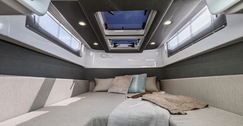 Cozy modern yacht cabin berth with twin skylight hatches, recessed LED lights, side windows with blinds, neutral cushions and a folded throw blanket