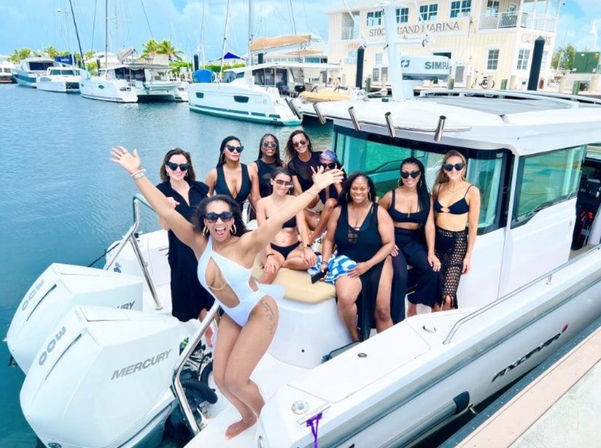 Fun group of women in swimsuits cheering on a white motorboat docked at a sunny marina with yachts and palm trees in the background.