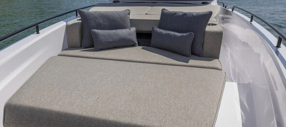 Bow sunpad on a motorboat with beige-gray upholstered cushions and matching lumbar pillows, black railings and calm water in the background