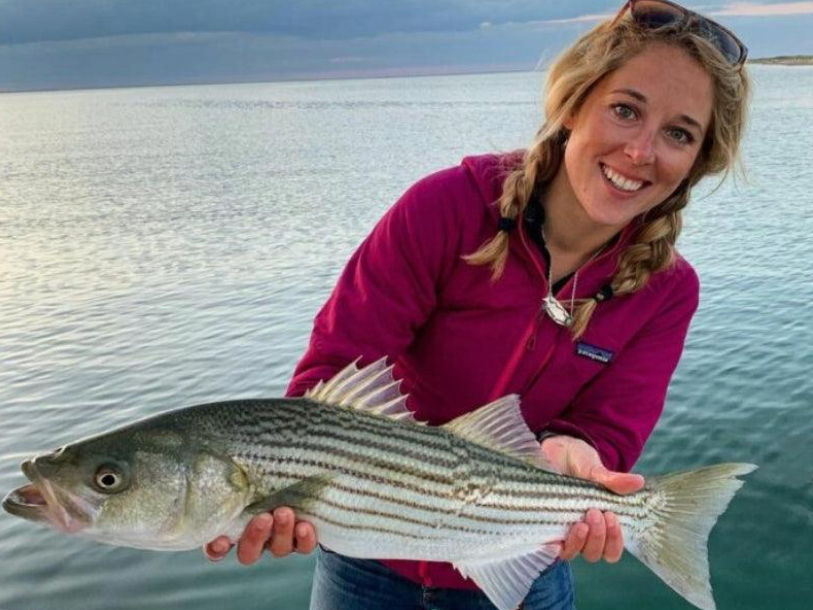 Smiling angler in a magenta jacket holding a striped bass aboard a boat over calm coastal waters at sunset — recreational saltwater fishing catch.