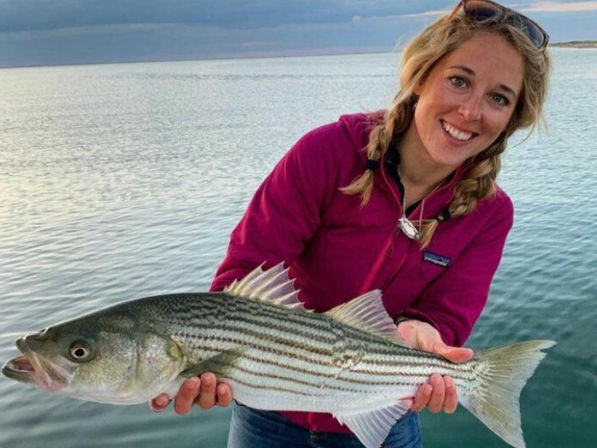 Smiling angler in a magenta jacket holding a striped bass aboard a boat over calm coastal waters at sunset — recreational saltwater fishing catch.