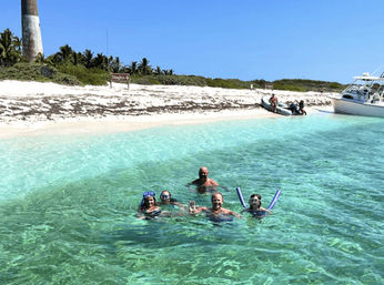 Smiling snorkelers bobbing in crystal-clear turquoise water near a white-sand tropical island beach with palm trees, a lighthouse, dinghies and an anchored boat.
