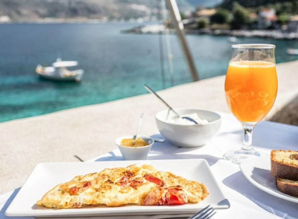 Sunny seaside breakfast on a terrace: tomato omelette on a white plate, toast, jam and yogurt bowls, glass of orange juice, turquoise bay with anchored boat in the background.