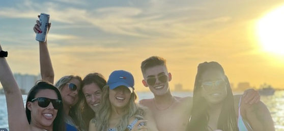 Smiling group of friends on a boat at golden sunset, wearing sunglasses and summer outfits, holding drinks with a city skyline and shimmering water in the background.