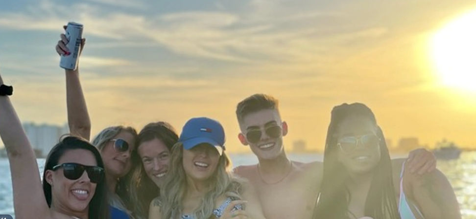 Smiling group of friends on a boat at golden sunset over the water, wearing sunglasses and a cap and holding drinks — summer coastal party vibe