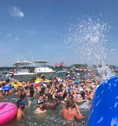 Crowded coastal boat party with dozens of people on inflatables and in shallow water near anchored motorboats, big water splash under a sunny blue sky