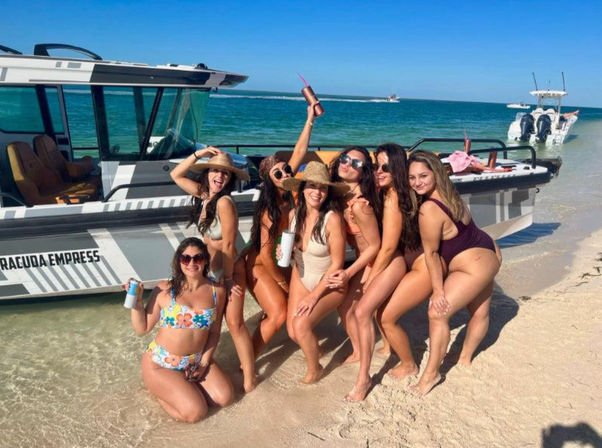 Seven women in colorful swimsuits smiling and posing with drinks on a sandy tropical beach beside a moored motorboat under a clear blue sky