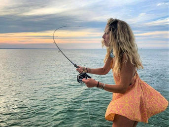 Angler casting a fly rod from a boat over calm coastal waters at sunset, wearing a flowing orange summer dress and long wavy blonde hair.