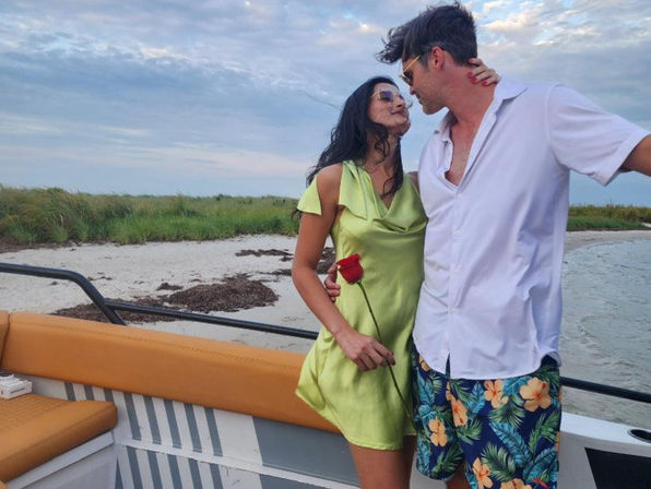 Romantic couple on a boat by a sandy shoreline and grassy dunes, woman in a lime-green dress holding a single red rose and man in a white shirt with tropical shorts leaning in under a cloudy sky