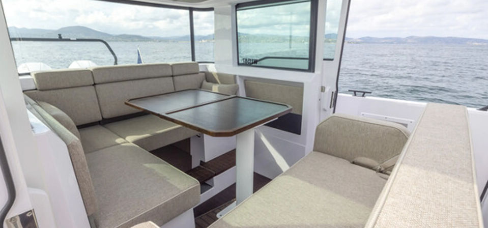 Sunlit motorboat cabin with beige U-shaped seating and a folding wood-trim table, large windows framing calm sea and distant coastal hills.