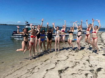 Group of friends in colorful swimsuits jumping and laughing on a sandy bay beach beside anchored boats under a bright blue summer sky.