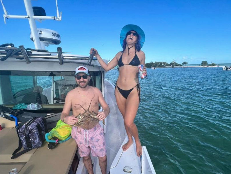 Two people on a boat in clear tropical waters — man holding a spiny lobster, woman in a black bikini and blue sun hat laughing with a canned drink, small island on the horizon.