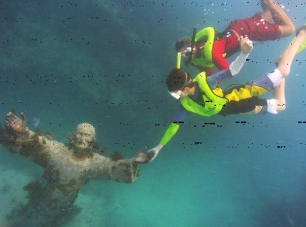 Two snorkelers in bright life vests and snorkel masks reach to touch an algae-covered underwater statue of Jesus in clear turquoise reef water