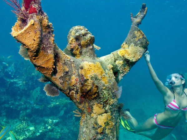 Snorkeler in a pink striped bikini reaches toward a coral-encrusted underwater statue with outstretched arms amid a tropical reef and clear blue water