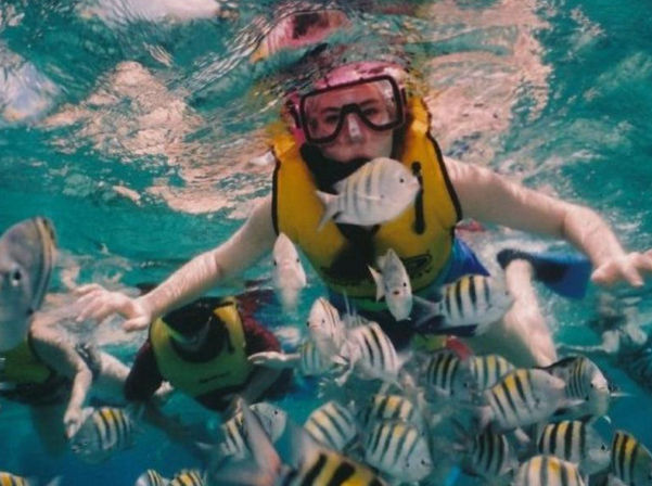 Snorkeler wearing a yellow life vest and pink mask swimming among a school of black-and-white striped sergeant major fish in clear turquoise tropical water