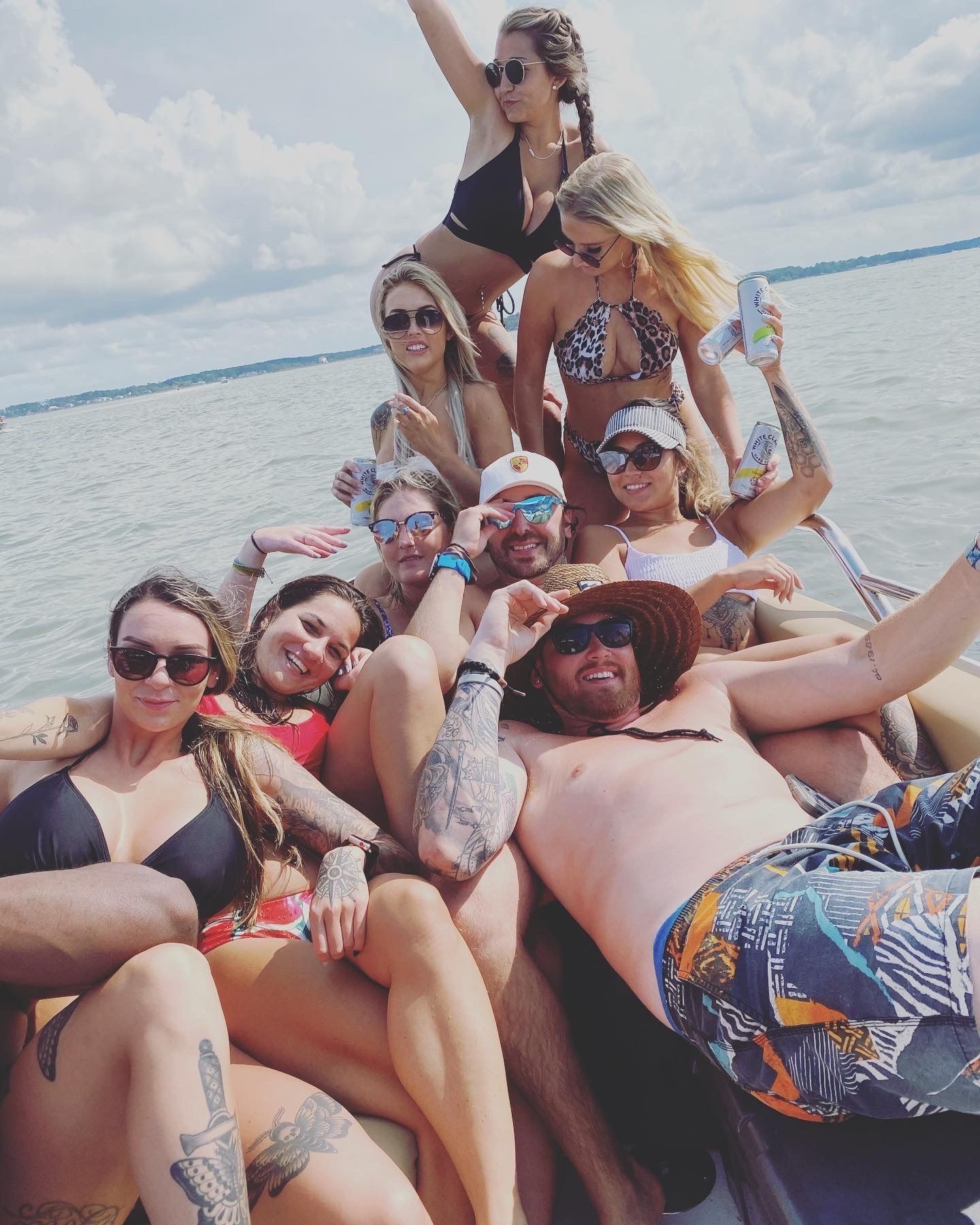 Group of friends on a lake boat enjoying a summer boat party, lounging in swimsuits and sunglasses with tattoos and canned drinks under a partly cloudy sky