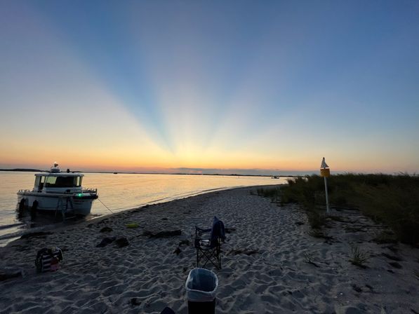 Beach sunset over a calm coastal inlet with crepuscular rays, a small motorboat anchored at the sandy shore, dunes and a folding chair on the beach.