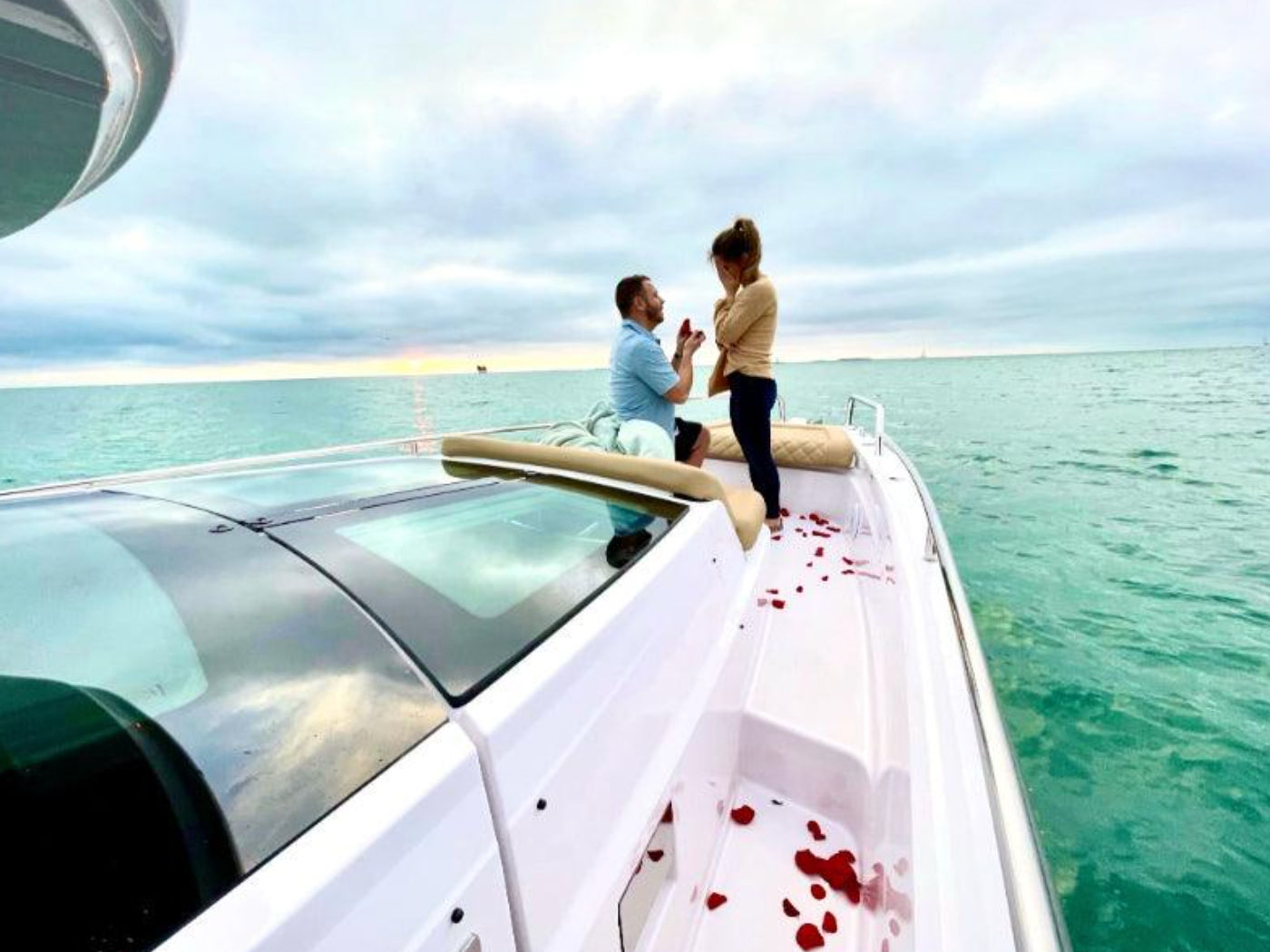 Romantic boat proposal on turquoise sea at sunset — man kneeling with a ring on the bow of a white motorboat, woman covering her face, rose petals scattered on the deck.