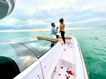 Romantic proposal on the bow of a white yacht — man kneeling with a ring as a woman covers her face, rose petals scattered on the deck, turquoise ocean and cloudy sunset horizon.