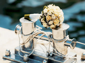 Cream and white rose bridal bouquet resting on a polished stainless-steel boat cleat on a sunlit dock, coiled ropes and blurred marina water in the background