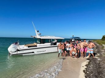 Cheerful group of people posing on a sandy beach beside a white motorboat anchored in shallow turquoise water under a clear blue sky