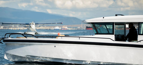 White commuter boat with silhouetted pilot in foreground as a seaplane lands on a coastal harbor, cargo ships and blue mountains in the background.