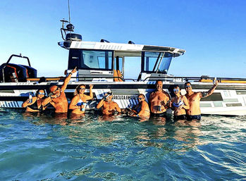 Smiling group of friends waist-deep in turquoise coastal water beside a modern cabin boat, holding drinks and taking selfies on a sunny boating party.