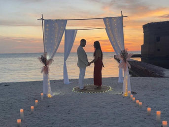 Silhouetted couple holding hands beneath a white draped wedding arch on a sandy beach at sunset, candles and fairy lights lining the oceanfront aisle.