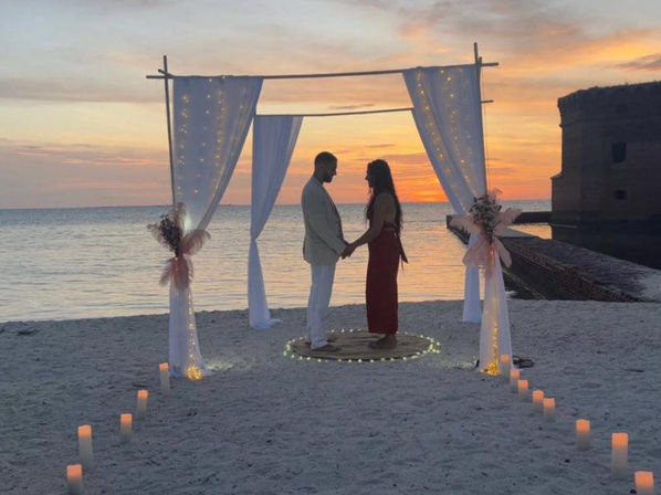Silhouetted couple holding hands beneath a white draped wedding arch on a sandy beach at sunset, candles and fairy lights lining the oceanfront aisle.