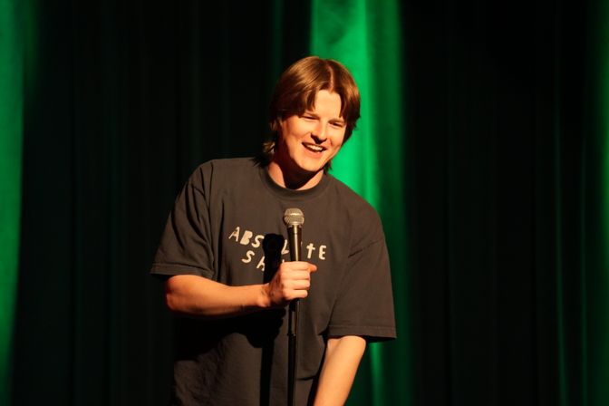 Stand-up comedian on stage smiling while holding a microphone, wearing a casual T-shirt with warm spotlight and green stage curtains in the background