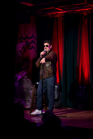 Stand-up comedian on a small comedy club stage with red curtains, holding a microphone and pointing at the audience while wearing sunglasses and a brown leather jacket; stool and a mounted bear prop visible in the dimly lit venue.
