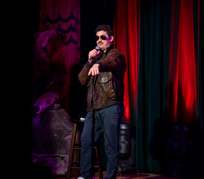 Stand-up comedian on a small comedy club stage with red curtains, holding a microphone and pointing at the audience while wearing sunglasses and a brown leather jacket; stool and a mounted bear prop visible in the dimly lit venue.