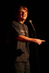 Young comedian on a dark comedy club stage, smiling and pointing while speaking into a microphone under a warm spotlight.