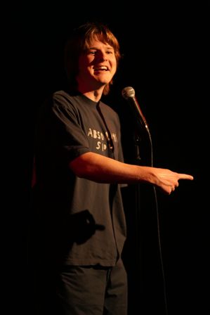 Young comedian on a dark comedy club stage, smiling and pointing while speaking into a microphone under a warm spotlight.