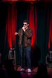 Stand-up comedian in sunglasses and brown leather jacket pointing at the audience while holding a microphone on an intimate comedy-club stage with red and green curtains, a stool with notes, and warm stage lighting.