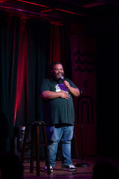 Stand-up comedian smiling into a microphone on an intimate comedy club stage, standing by a stool under red and purple stage lights and dark curtains.